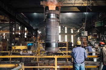 Tres operarios con cascos de seguridad observan un cilindro metálico incandescente suspendido por una grúa en un taller industrial