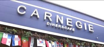 Fachada de un edificio azul con el letrero 'CARNEGIE DINER & CAFE' en letras blancas. Debajo, una hilera de banderas nacionales y follaje verde
