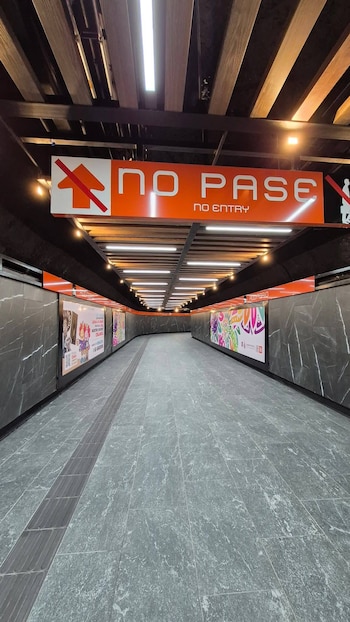 Corredor renovado y vacío de una estación de metro con paredes oscuras y un cartel naranja de "NO PASE" en el techo, iluminado por luces LED