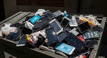 Contenedor de basura lleno de teléfonos celulares desechados, incluidos smartphones modernos con pantallas agrietadas y algunos con el logo de AT&T México visible.