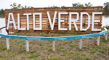 Cartel de madera con letras blancas que forman el texto ALTO VERDE, sobre un terreno de césped junto a una baranda azul y agua.