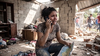 Una mujer llora mientras habla por teléfono celular y sostiene un mapa dentro de una habitación destruida con escombros y grietas.