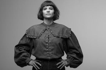 Retrato en blanco y negro de una mujer de cabello corto y flequillo, con una blusa de mezclilla con cuello de volantes y cordones, manos en la cintura