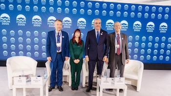 Cuatro personas están de pie ante un fondo azul con la frase Forum 2000, delante de sillones blancos y mesas con identificación