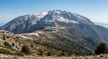 Montaña griega con cumbres nevadas y rocosas bajo cielo azul claro. Laderas inferiores muestran parches de nieve derretida, vegetación mediterránea y suelo seco.