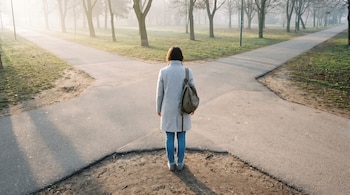 Persona de espaldas con abrigo gris y mochila en cruce de caminos de asfalto y tierra en parque. Árboles sin hojas y neblina de fondo con sol de amanecer.