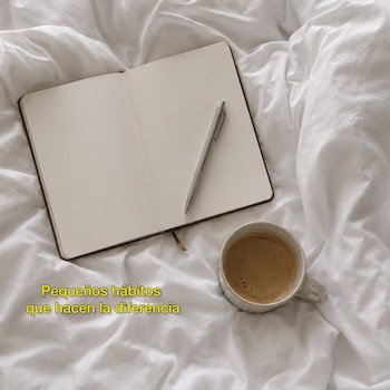 Un cuaderno abierto con un bolígrafo plateado sobre páginas en blanco, junto a una taza de café en sábanas blancas arrugadas