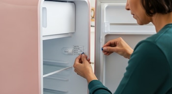 Primer plano de las manos de una persona adulta que apunta a una etiqueta de especificaciones técnicas dentro de un refrigerador rosa abierto.