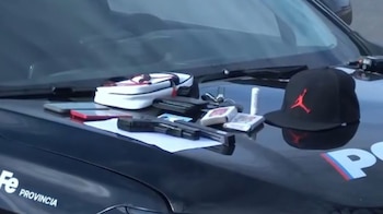 Una réplica de arma de fuego, dos teléfonos, una cartera, una bolsa y una gorra negra con logo rojo sobre el capó oscuro de un vehículo policial con el texto "PROVINCIA"