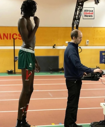 Un hombre alto con dreadlocks y shorts verdes tiene sensores adheridos a su cuerpo. Un técnico rubio usa un teclado y ratón frente a él en una pista de atletismo