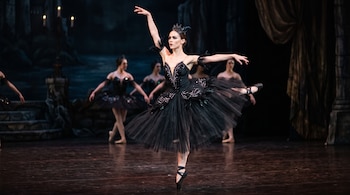 Una bailarina con tutú negro se sostiene en puntas sobre un escenario junto a otras bailarinas de fondo frente a una escenografía.