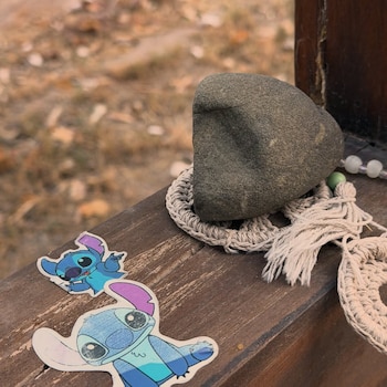 Una piedra oscura descansa sobre un adorno de ganchillo, junto a dos calcomanías de Stitch y cuentas en una superficie de madera, con follaje borroso detrás