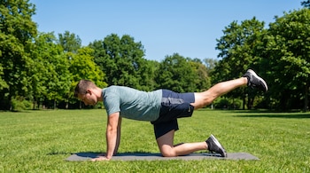 Un hombre en ropa deportiva sobre una colchoneta de ejercicio en el césped de un parque, con una pierna extendida hacia atrás, árboles de fondo y cielo azul.