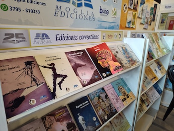 Estante con libros de diferentes portadas, títulos y colores. En la parte superior hay carteles con el logo y nombre de Moglia Ediciones y el teléfono 3795-810333