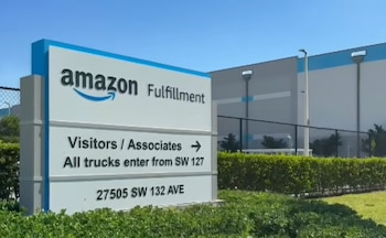 Un letrero azul y blanco de Amazon Fulfillment frente a un gran almacén gris bajo un cielo azul claro, con vegetación y una valla negra en primer plano