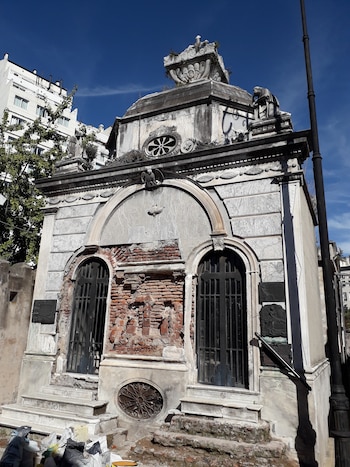 Mausoleo de piedra deteriorado con dos puertas de rejas metálicas, ladrillos expuestos, detalles tallados, edificio blanco, árbol y poste de luz