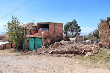 Vista exterior de una casa parcialmente destruida, con muros de ladrillo expuestos, escombros de tierra y ladrillo en el suelo, dos puertas verdes y un cielo azul claro