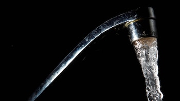 Un grifo de metal plateado en primer plano, inclinado hacia la derecha, con un chorro constante de agua clara cayendo hacia abajo sobre un fondo negro