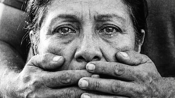 Fotografía en blanco y negro del primer plano de una mujer latina. Múltiples manos cubren su boca. Tiene lágrimas en los ojos y mira fijamente.