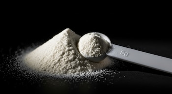 Fotografía macro de un montículo de polvo de creatina blanco junto a una cuchara dosificadora con "5g" sobre un fondo negro, con luz lateral.