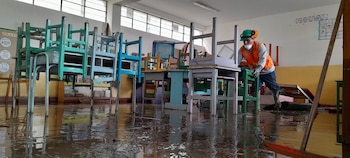 Un aula escolar inundada con mobiliario apilado, mesas y sillas. Un trabajador con equipo de protección limpia el agua del piso