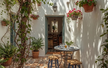 Vista de un patio soleado con paredes blancas, plantas trepadoras, macetas y flores colgantes. Una mesa redonda con dos taburetes y servicio de mesa para dos. Se ve una puerta abierta al interior.