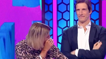 En un plató de televisión vibrante, una mujer rubia con blusa estampada se cubre la boca, mientras un hombre sonriente con chaqueta oscura y camisa blanca mira al frente