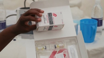 Mano sosteniendo una caja de Lenacapavir 300 mg. En primer plano, una caja abierta con viales. Al fondo, un vaso azul y una botella de agua