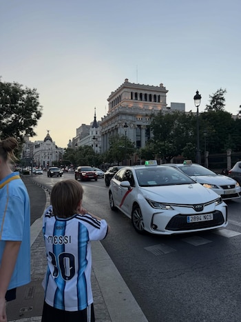 Un niño de espaldas con camiseta de fútbol de Messi número 30 en la acera, mirando coches y taxis en una calle de la ciudad con edificios al fondo