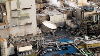 Vista aérea de una planta industrial con un gran tanque de almacenamiento blanco colapsado, rodeado de edificios, tuberías y una zona inundada