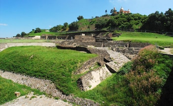 Restos arqueológicos de piedra, escalinatas y montículos de tierra con pasto verde; en el fondo, una colina con un santuario de cúpulas anaranjadas