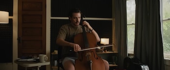 Un hombre (Pedro Pascal) sentado toca un violonchelo con un arco. Lleva camiseta y pantalón corto. Hay paneles acústicos, una puerta, una ventana y un escritorio