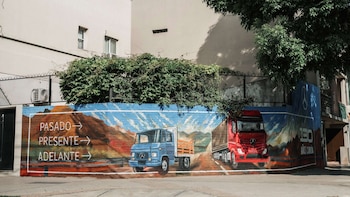 Mural en una pared exterior con dos camiones pintados, uno azul y otro rojo, situado bajo una hilera de arbustos junto a la acera