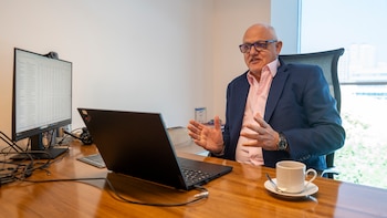 Javier Timerman con gafas y chaqueta azul gesticula junto a una computadora portátil, un monitor y una taza de café