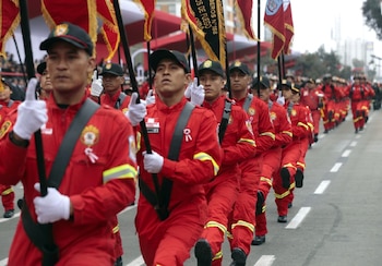 Fila de bomberos hombres uniformados de rojo con gorras negras y guantes blancos marchando. Sostienen banderas rojas, amarillas y negras en un desfile al aire libre