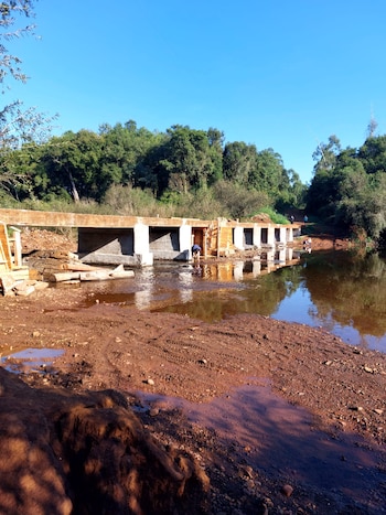 Un puente de concreto en construcción cruza un río de agua marrón y orillas fangosas, rodeado de frondosos árboles verdes bajo un cielo azul claro