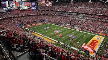 Vista aérea de un estadio de fútbol americano lleno de espectadores, con el campo verde, pantallas gigantes mostrando el Super Bowl LVII y humo rojo