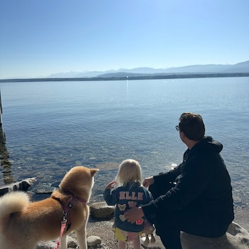 Un hombre, una niña de espaldas y un perro con arnés miran hacia un lago de aguas claras con montañas en el horizonte