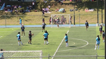 Múltiples jugadoras de fútbol femenino en un campo de césped artificial. Se ven dos porterías, una pista de atletismo azul y una colina con personas sentadas