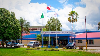 Edificio de la Universidad Científica del Perú con letrero frontal, tres banderas en mástiles, árboles, palmeras y varios autos estacionados