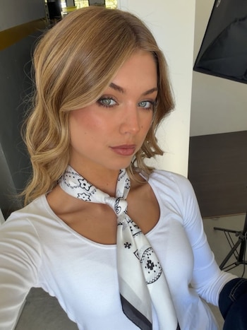 Mujer joven de cabello rubio y ojos claros, vestida con una camiseta blanca de manga larga y un pañuelo blanco con estampado floral negro atado al cuello