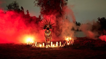 Una persona con un penacho de plumas se arrodilla en el centro de un círculo de velas encendidas rodeada por humo rojo