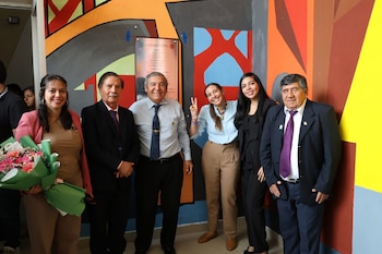 Seis personas posan sonriendo frente a una pared con murales coloridos y una placa. Una mujer sostiene un ramo de flores de colores
