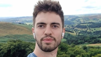 Un hombre joven con barba corta y auriculares inalámbricos blancos mira a la cámara frente a un paisaje de colinas verdes