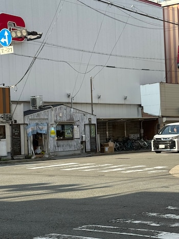 Calle con cruce peatonal, un pequeño edificio de madera con banderas, bicicletas estacionadas bajo un techo, y un SUV blanco en movimiento a la derecha