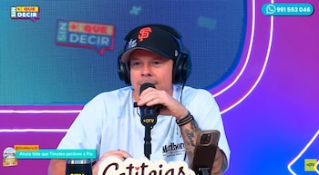 Hombre con gorra negra, auriculares y camiseta blanca, sentado. Habla por un micrófono negro. Tiene tatuajes en el brazo y pulseras. Un teléfono móvil está en la mesa