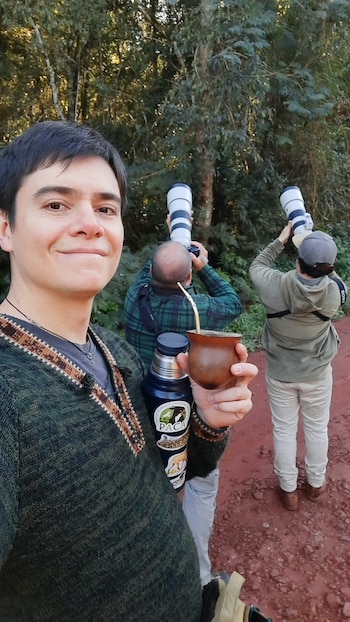 Un hombre sostiene un mate frente a dos personas que apuntan cámaras con teleobjetivo hacia los árboles en un camino de tierra rojiza