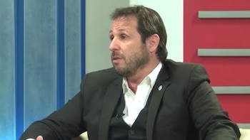 Hombre de mediana edad con barba y traje oscuro en un estudio de televisión. El fondo presenta paneles azules y rojos con detalles horizontales