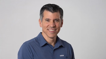 Un hombre de mediana edad, David Uzcátegui, con cabello oscuro y gris, sonríe mirando a la cámara, vestido con una camisa azul y con un fondo blanco liso