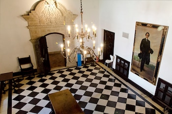 Interior de un salón con piso de baldosas blancas y negras, araña de bronce con velas, arco de piedra tallada, retrato de un hombre, dos puertas oscuras y sillas